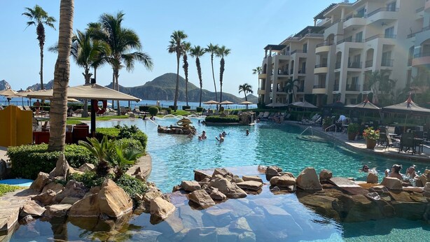Uma área de piscina em resort com água cristalina, palmeiras e uma montanha ao fundo.