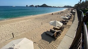 Uma praia com fileiras de espreguiçadeiras e guarda-sóis, vista clara do oceano e colinas ao longe.