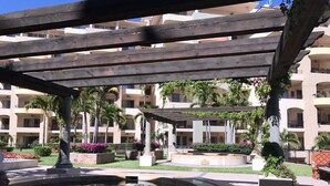 Um pátio com uma fonte central, rodeado por palmeiras e um edifício de vários andares com varandas.