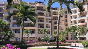 Um hotel de vários andares com varandas, palmeiras e um jardim bem cuidado com flores roxas.