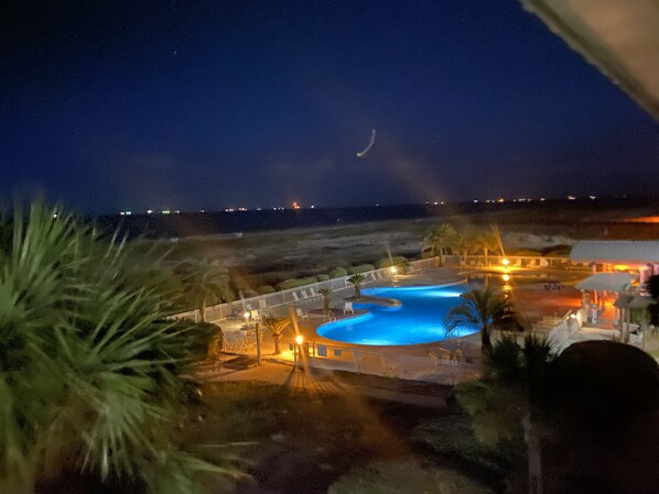 Une vue nocturne d’un complexe hôtelier avec une piscine, des palmiers et une plage en arrière-plan.