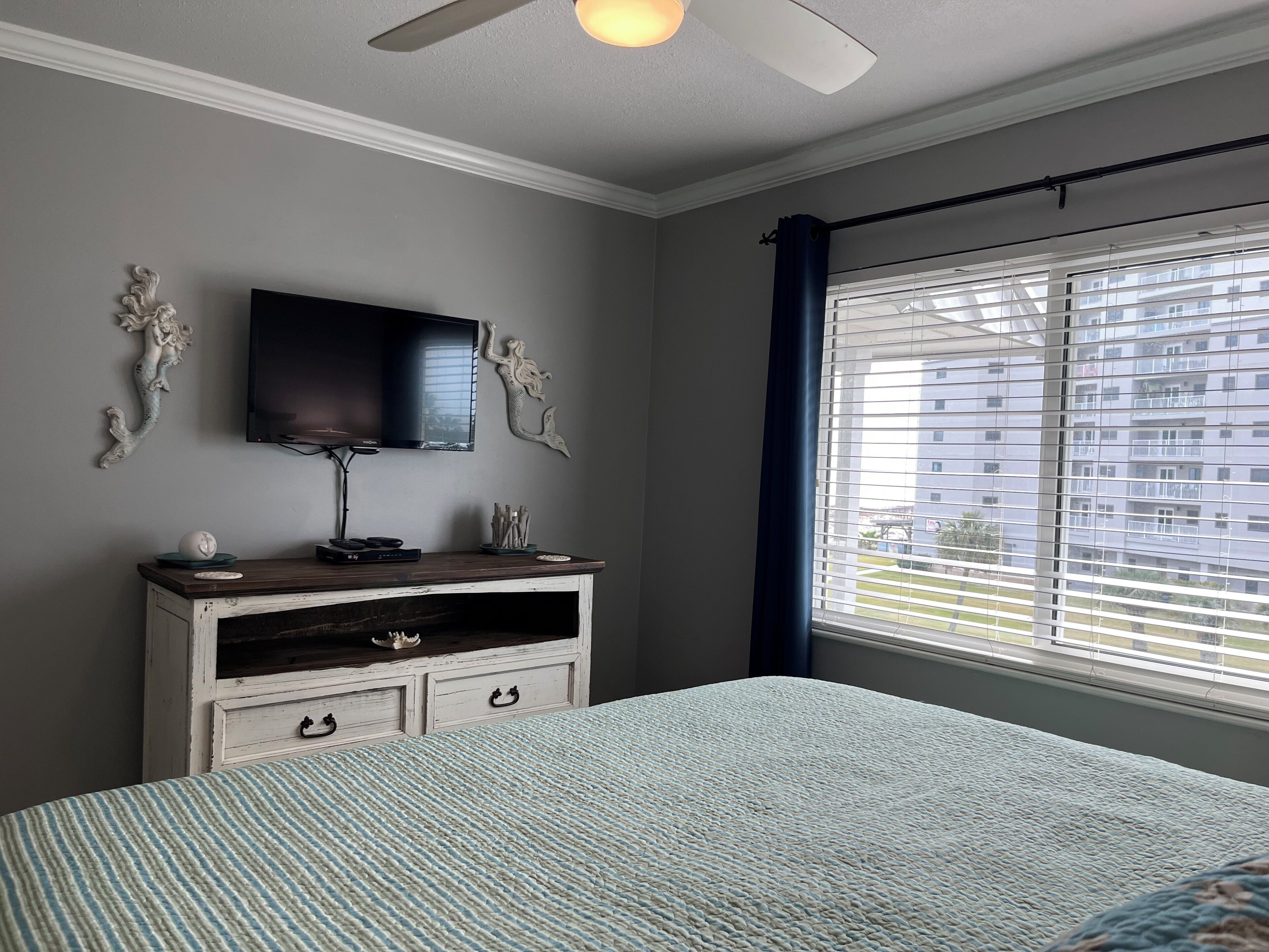 A bedroom with a bed, a TV mounted on the wall, a dresser, and a window with blinds.