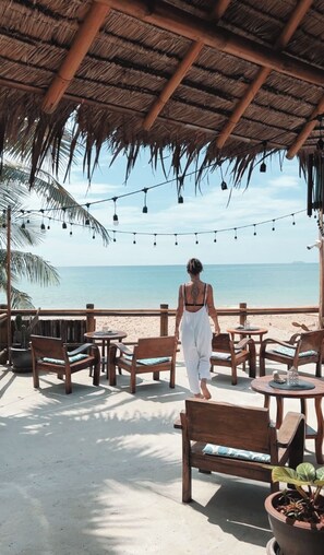 Un patio frente a la playa con muebles de madera, techo de paja e iluminación colgante.