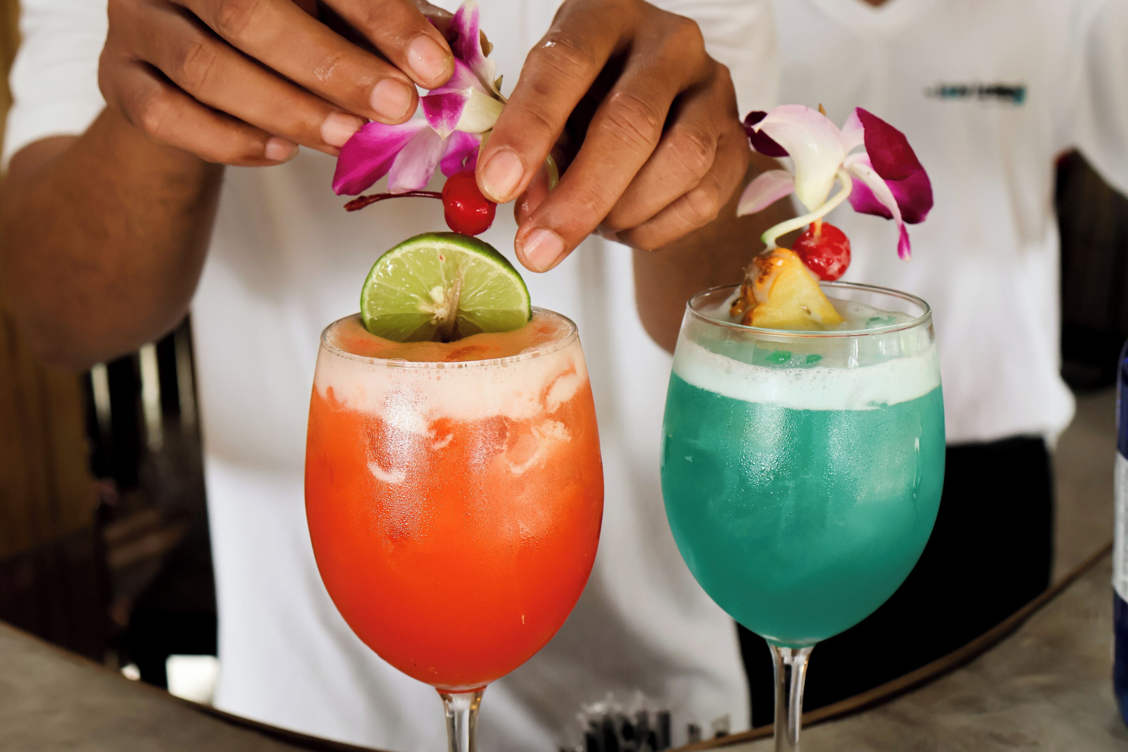 Two cocktails garnished with flowers and fruit, served in stemmed glasses.