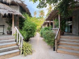 A pathway flanked by thatched-roof structures with bamboo railings, leading to a building with large windows and a view of palm trees.