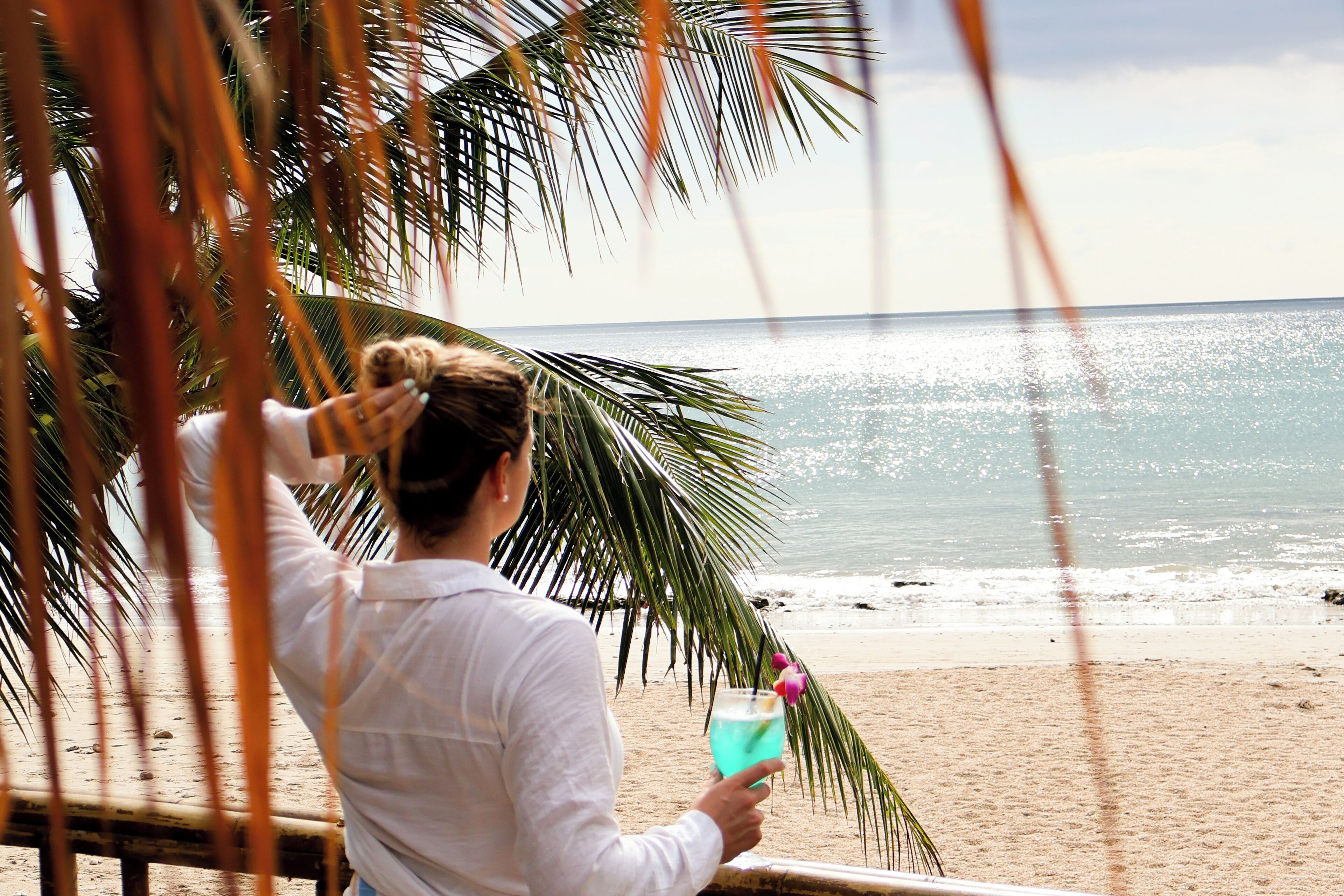 A person relaxing on a beach with a drink, palm trees, and the ocean in the background.