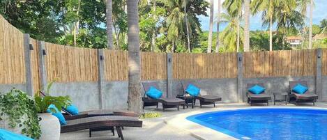 Zona junto a la piscina con sillones de descanso y una pequeña mesa, rodeada por una valla de bambú y palmeras.