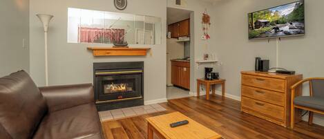 A living room with a fireplace, a sofa, a coffee table, a TV, and a kitchen in the background.