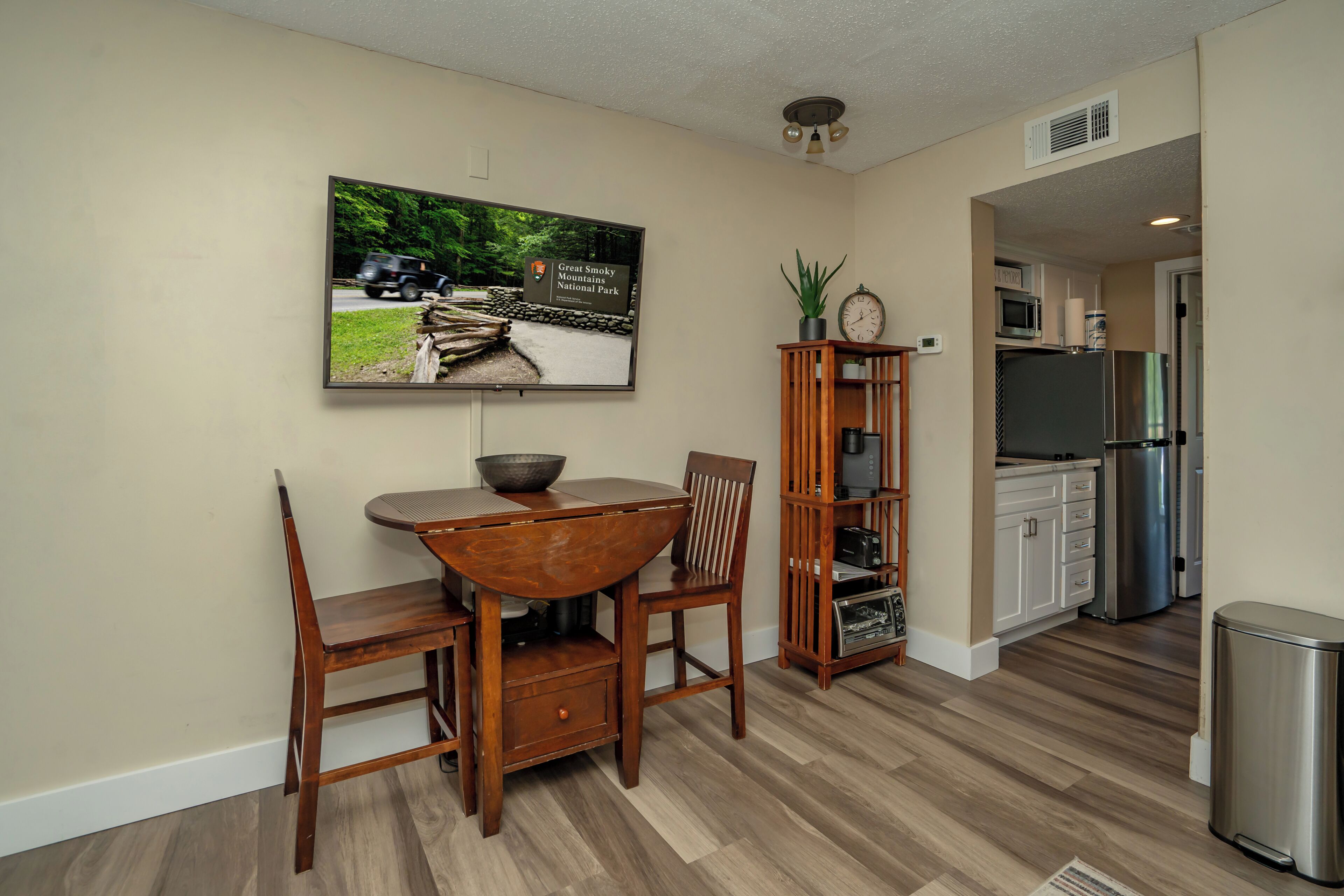 Un coin repas avec une table et des chaises en bois, un téléviseur fixé au mur et une cuisine en arrière-plan.