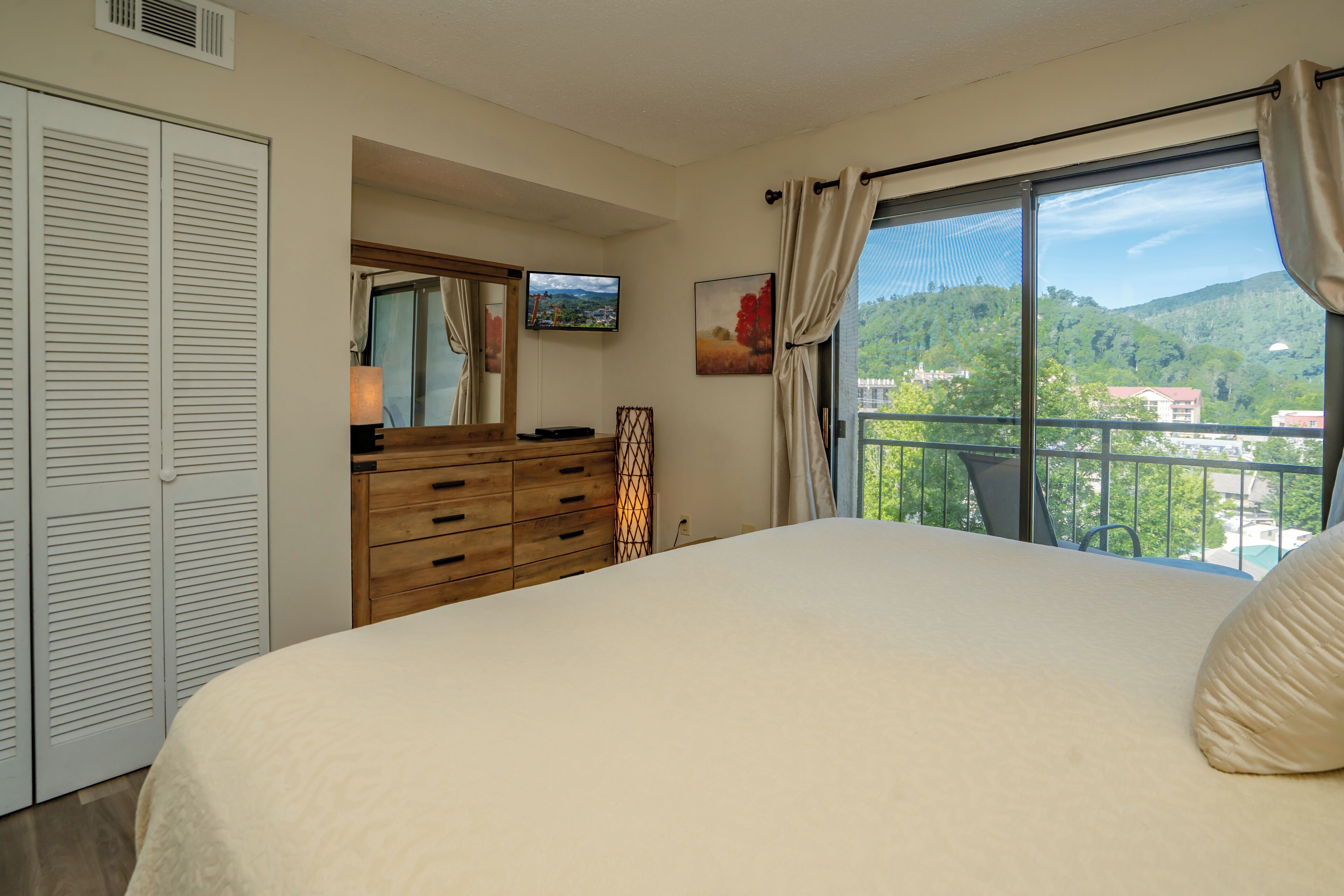 Ein Schlafzimmer mit einem großen Bett, einem Schminktisch, einem Fernseher und einem Balkon mit Blick auf Grünflächen.