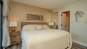 Un dormitorio con una cama grande, dos mesitas de noche con lámparas, un cabecero de madera, un espejo y una puerta que da a otra habitación.