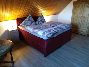 A bedroom with a wooden ceiling, a bed with patterned bedding, a nightstand, a wooden cabinet, and a round table.
