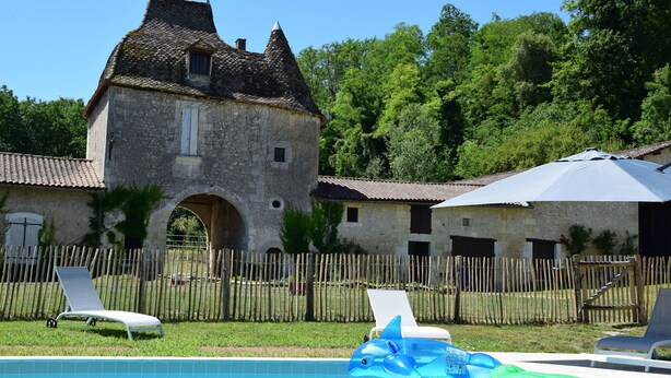 Una piscina con sillas de descanso y juguetes inflables, rodeada por una cerca de madera y un edificio de piedra con tejado de azulejos.
