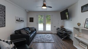 Ein modernes Wohnzimmer mit einem schwarzen Ledersofa, einem grauen Teppich, einem Deckenventilator, einem Flachbildfernseher und einem Regal mit einem 'Namaste'-Schild.