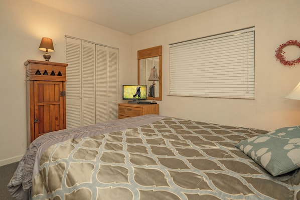 Un dormitorio con cama, televisor sobre un aparador, armario y ventana con persianas.