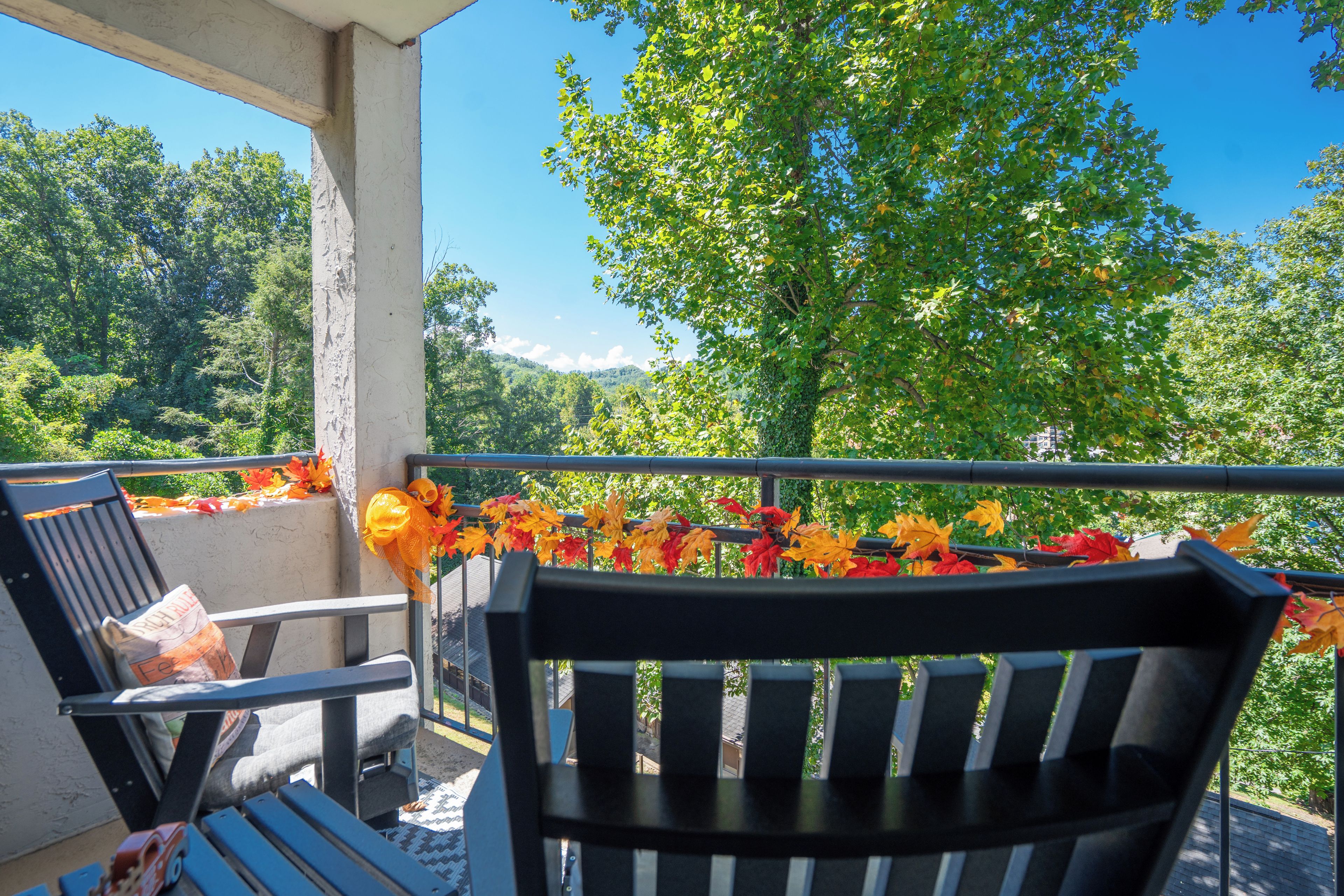 A balcony with two chairs and a table, overlooking a forested area with autumn leaves.