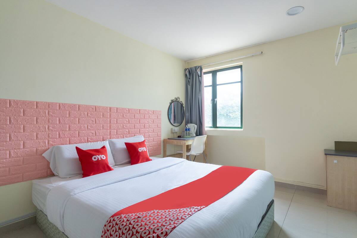 A hotel room with a brick-patterned headboard, a bed with white bedding and red accents, a desk with a chair, and a window with curtains.