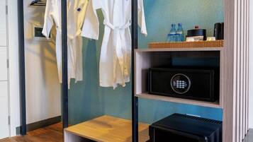 Una habitación de hotel moderna con suelo de madera, un perchero de metal negro con batas blancas, un estante con artículos de aseo y un dispositivo electrónico negro.