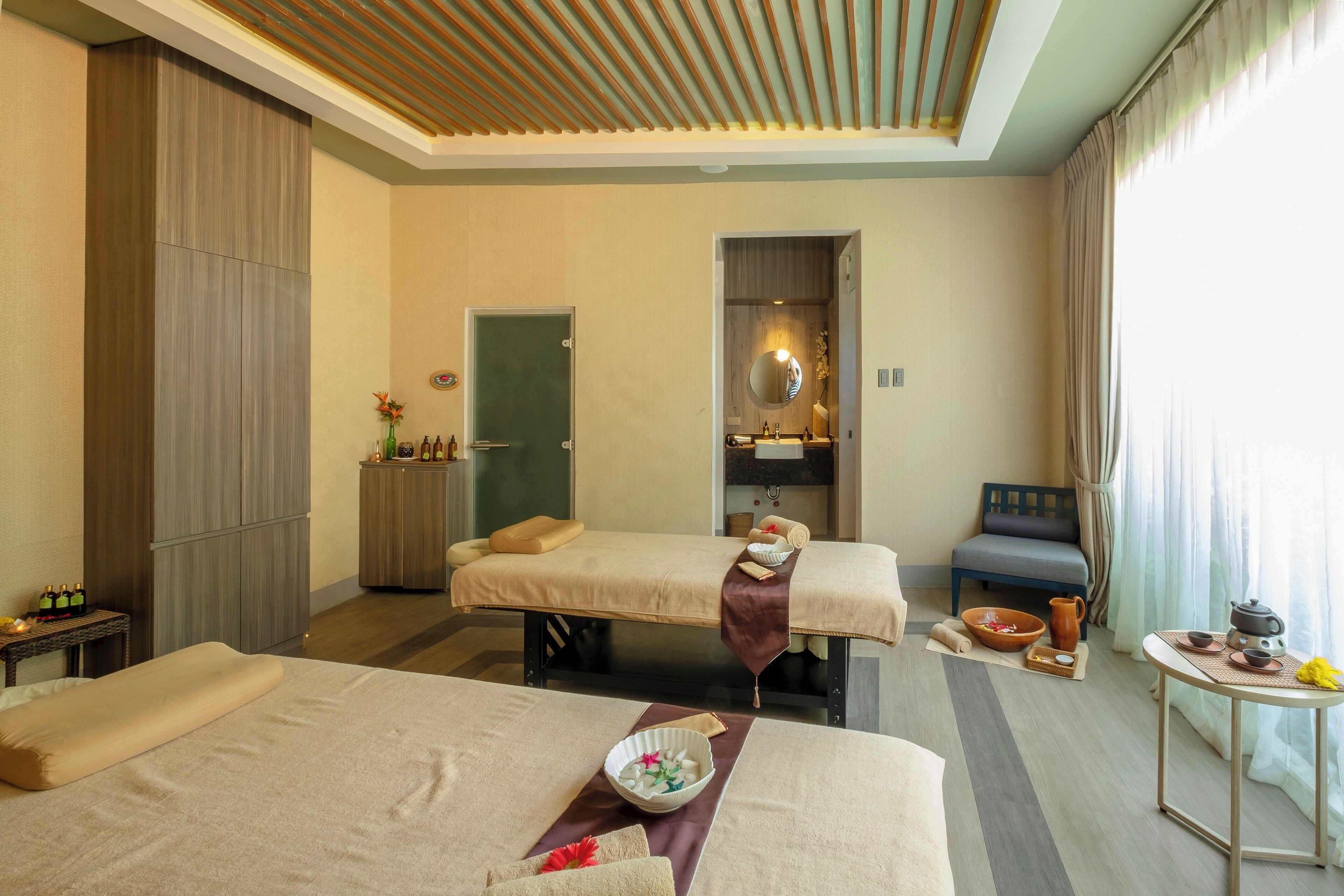 Habitación de hotel con dos camas, una camilla de masaje, una mesita con juego de té y vistas al exterior.