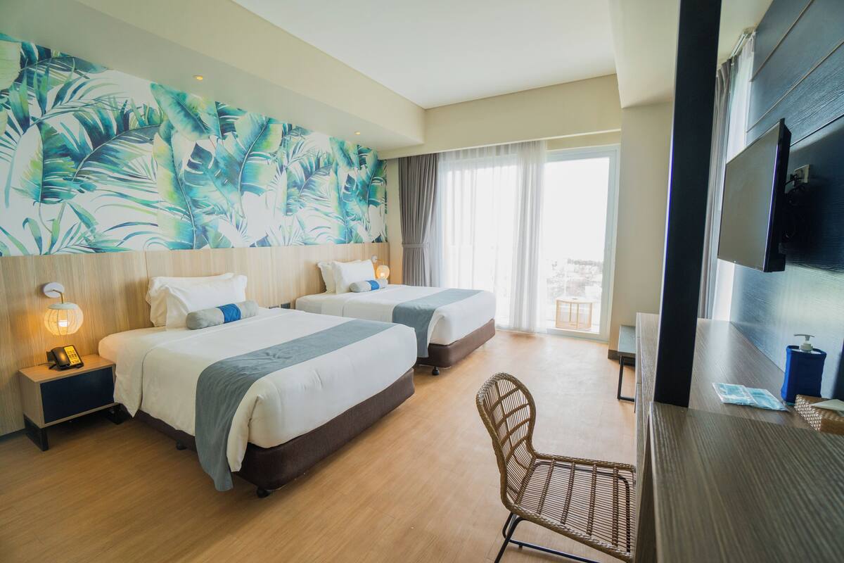 A hotel room with two beds, a large window with a view, a TV, and a wall with a tropical leaf mural.