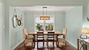 Un comedor con una mesa de madera, cuatro sillas de mimbre, una lámpara colgante y un ventanal con plantas.