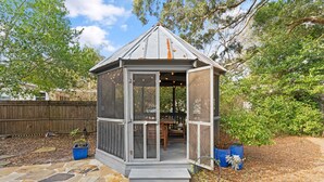 Una pequeña estructura exterior cerrada con techo de metal y paredes de vidrio, rodeada por un patio de piedra y valla de madera.