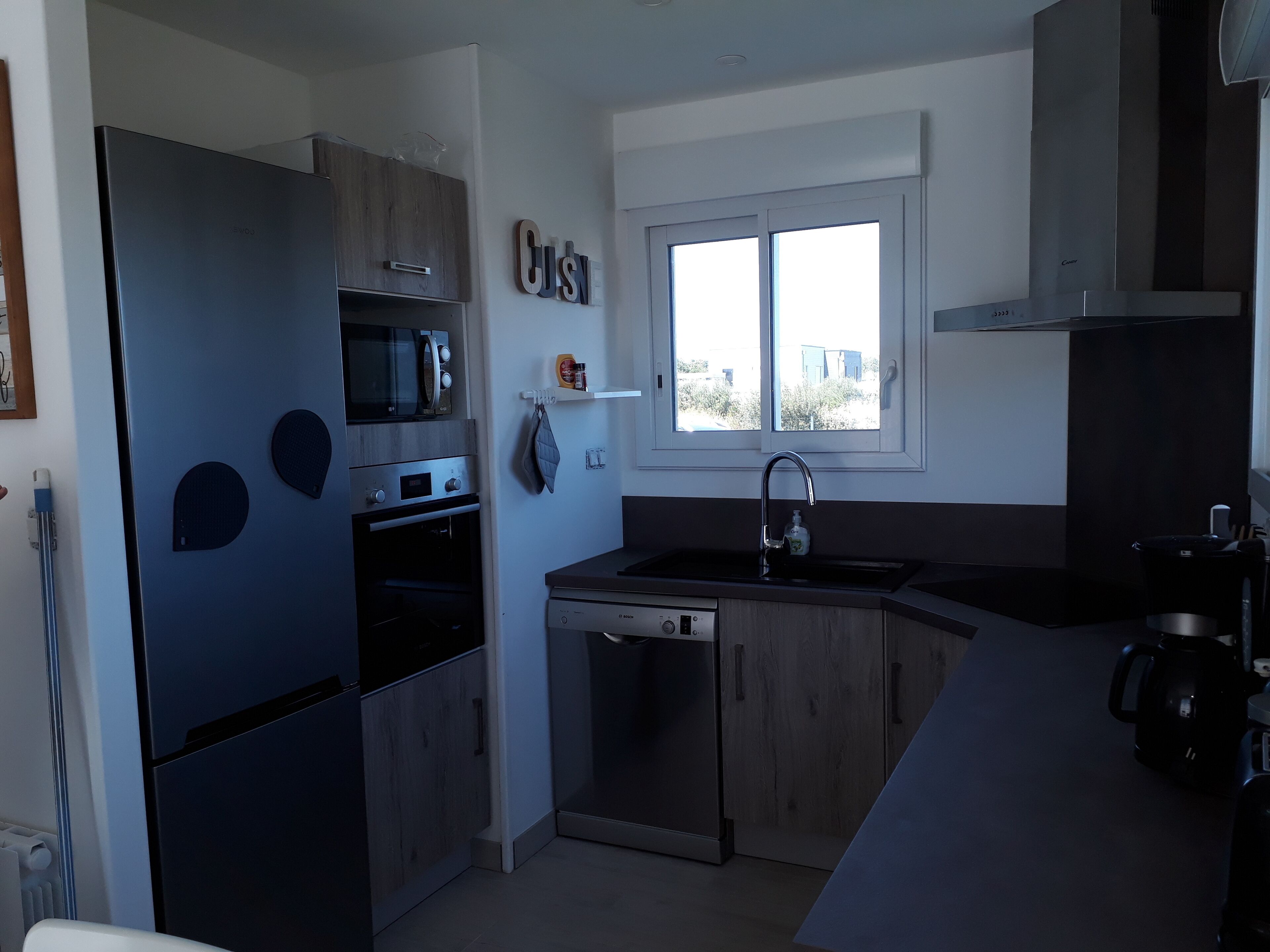 Eine moderne Küche mit Kühlschrank, Mikrowelle, Backofen und Geschirrspüler. Über der Spüle befindet sich ein Fenster und eine Arbeitsplatte mit einem Wasserkocher.