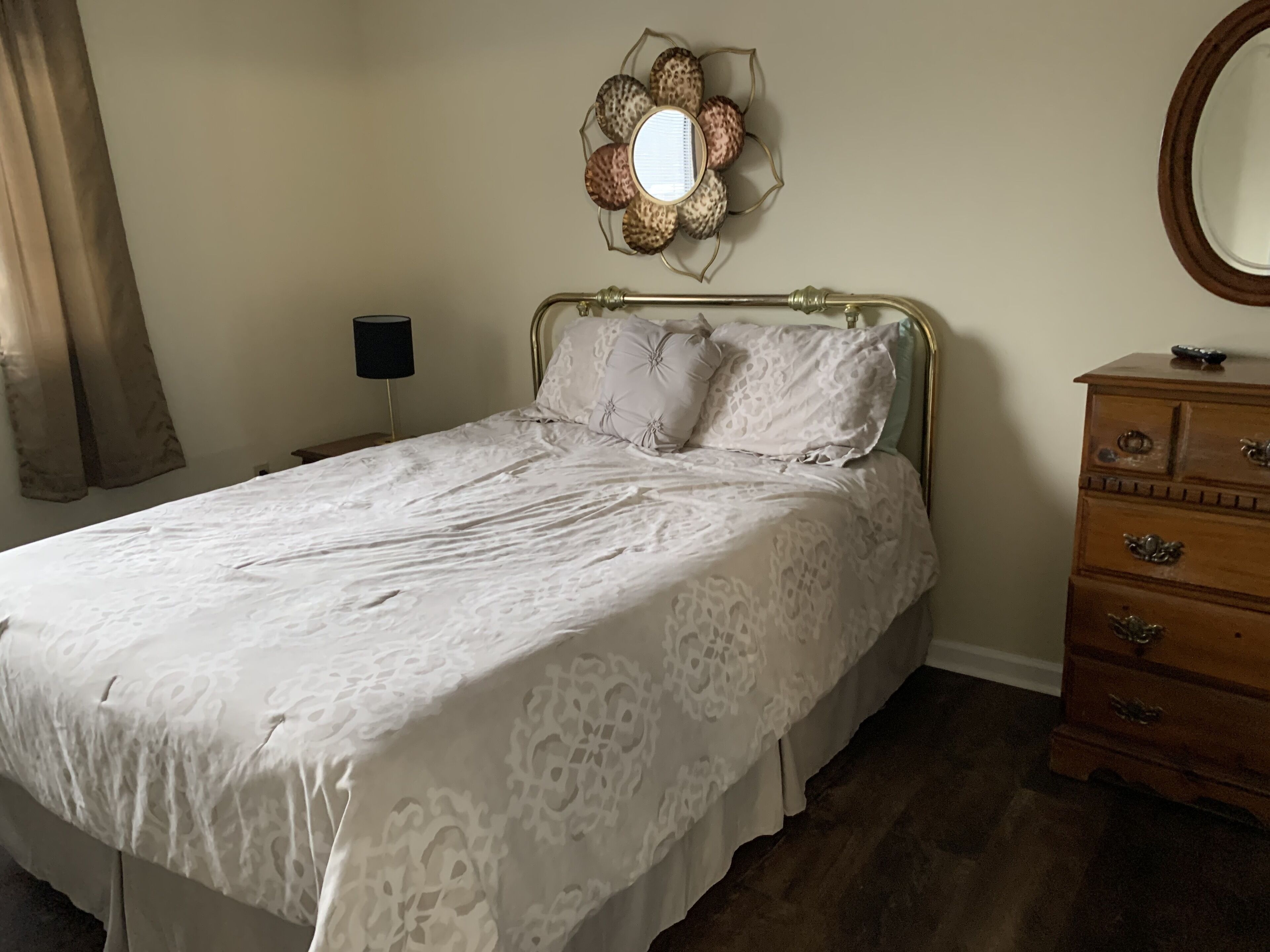 Uma cama bem arrumada com colcha estampada, cômoda de madeira com gavetas e espelho fixado na parede.