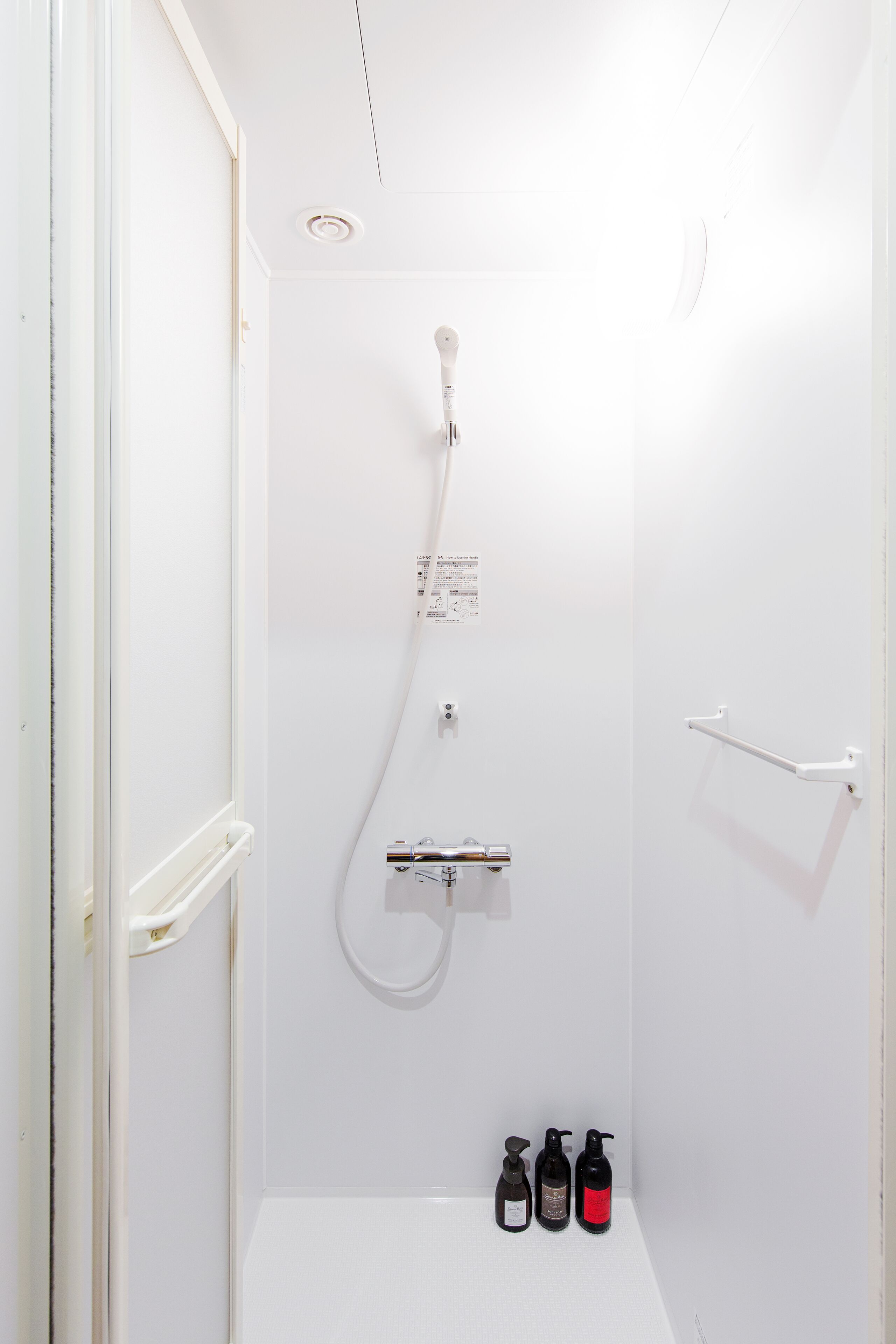 Un baño pequeño y blanco con cabezal de ducha manual, un perchero y dos botellas de champú y acondicionador.