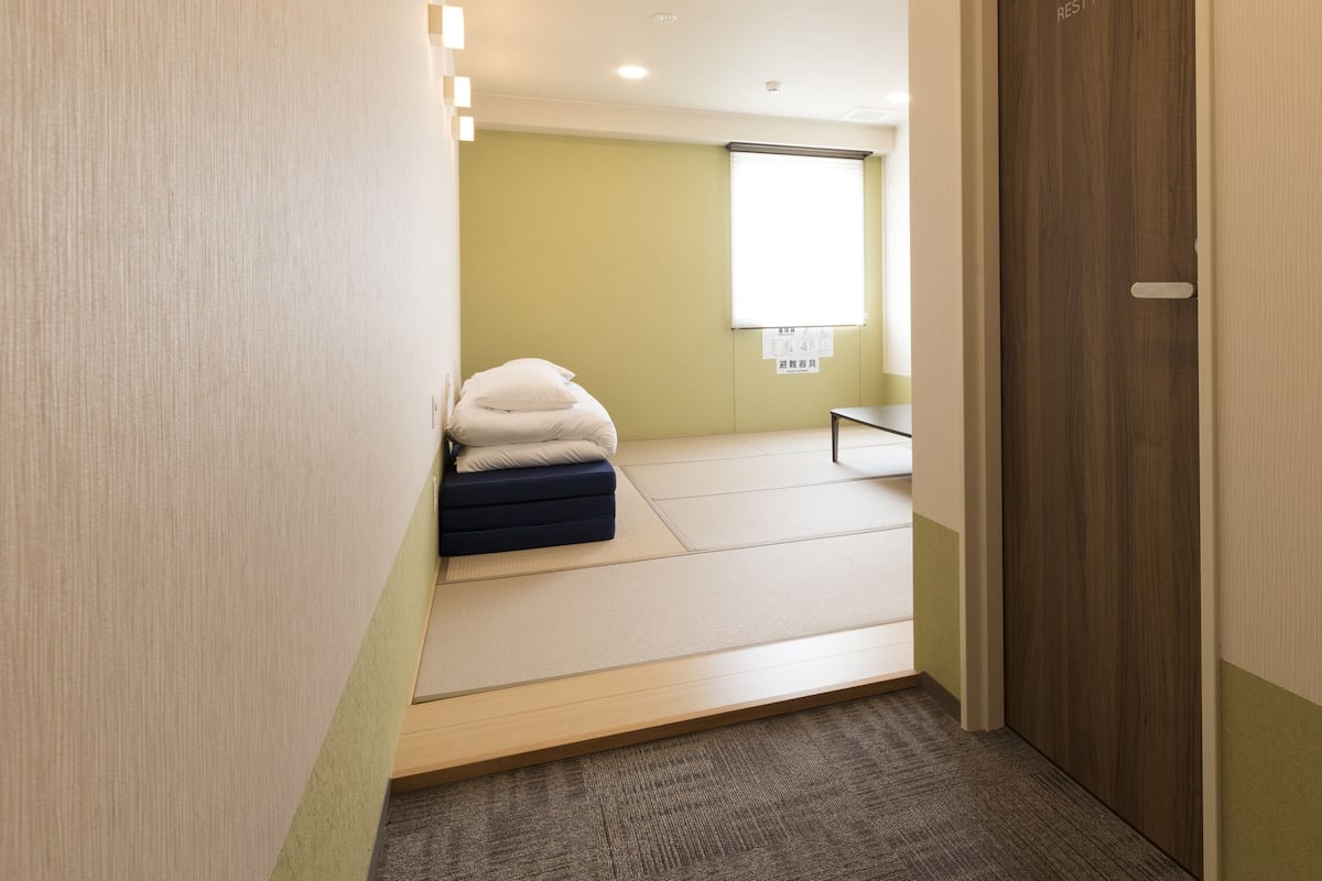 A room with tatami mat flooring, a bed with a white and blue mattress, a small table, and a window with a white blind.