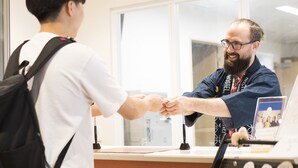 Two individuals shaking hands at a reception desk with a laptop and a framed photo on the counter.