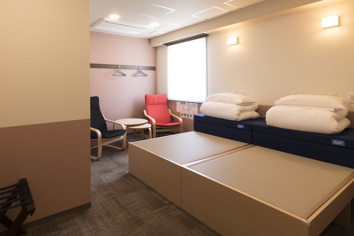 A hotel room with a bed, a red chair, a small table, and a window with blinds.