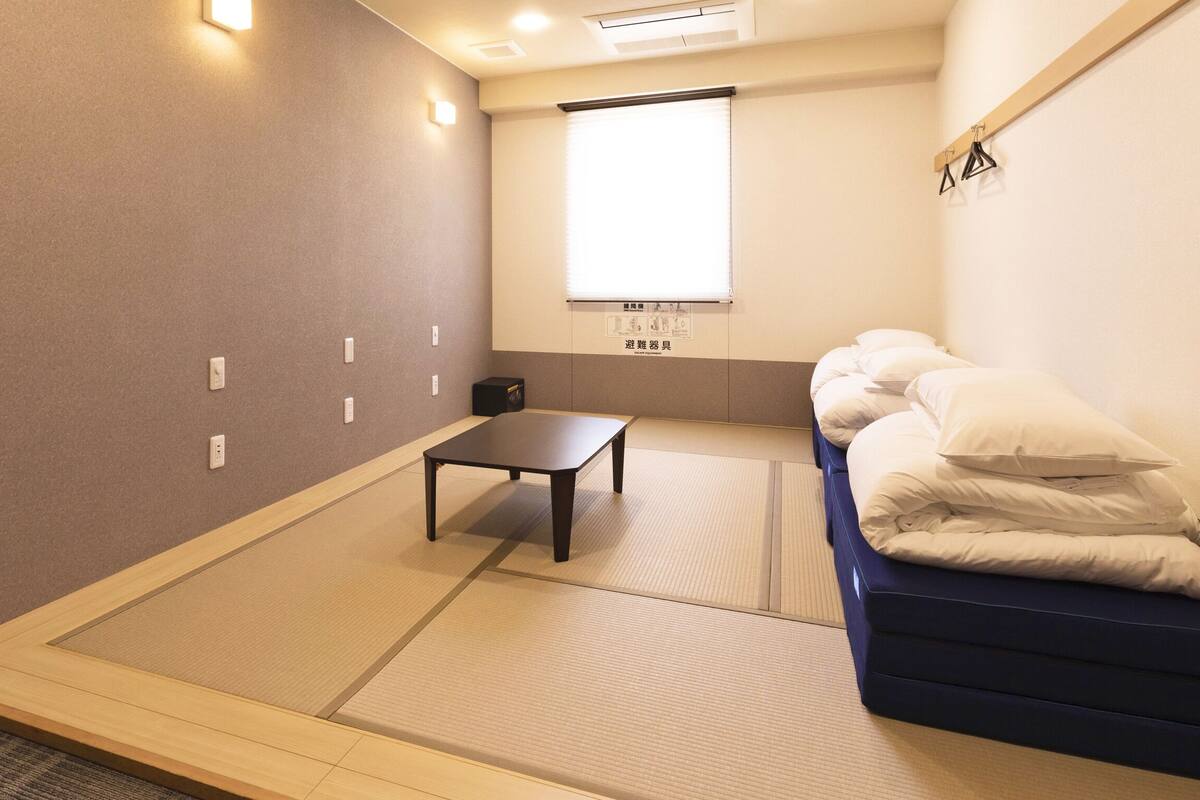 A room with tatami flooring, a low table, a bed with white bedding, and a window with blinds.