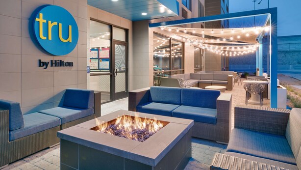 Een buitenzitgedeelte met banken en een vuurkuil, een blauw bord met 'tru by Hilton' en lichtjes.