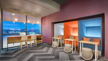A modern hotel room with a large window, a desk with two monitors, and a seating area with ottomans and a sofa.