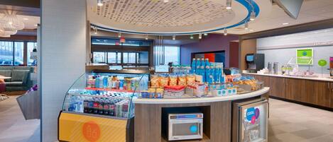 A modern self-service kiosk with a variety of beverages and snacks, a microwave, and a seating area with a view of the city.