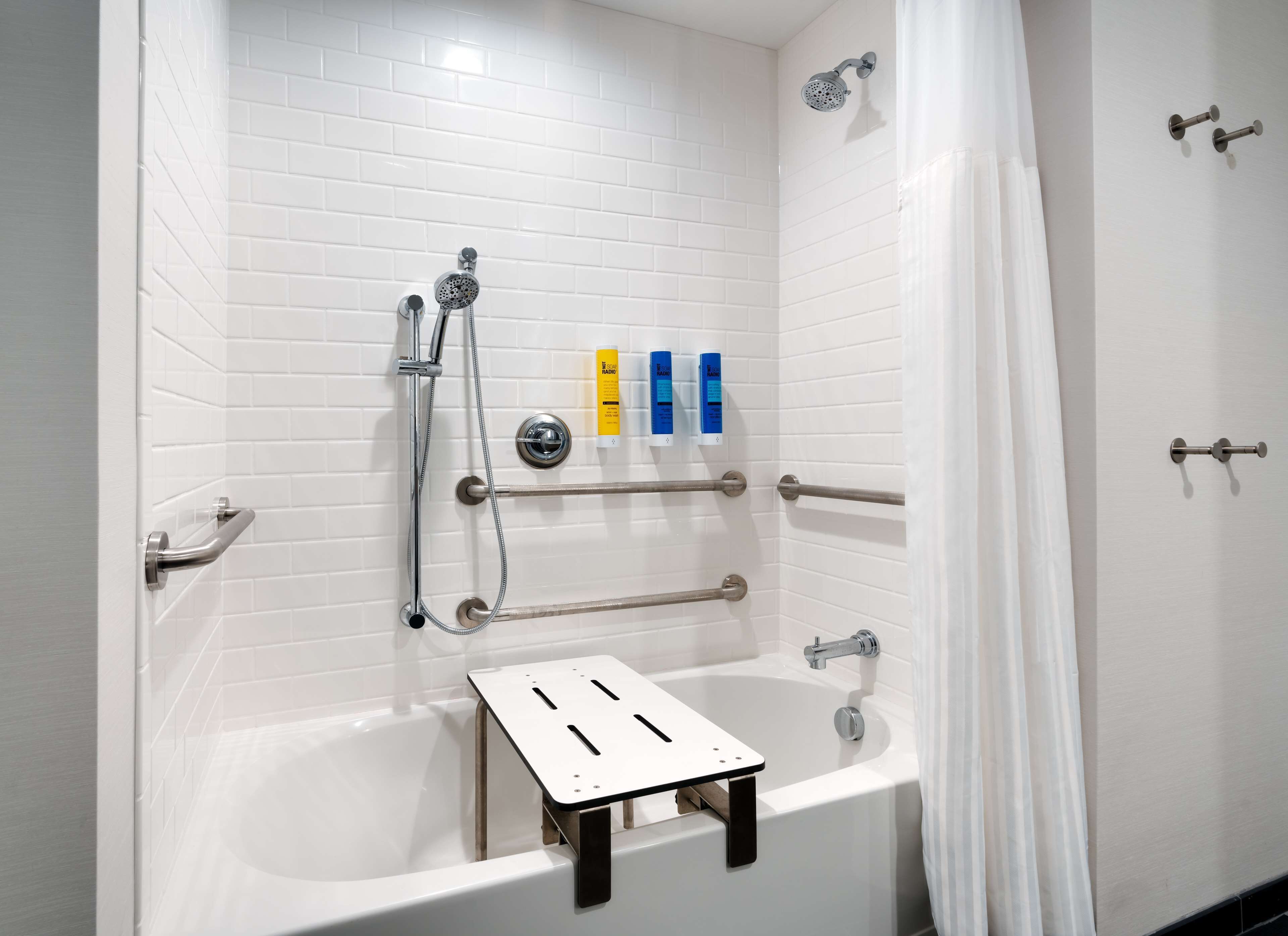 A modern bathroom with white tiled walls, a bathtub, a showerhead, grab bars, and a small shelf with toiletries.