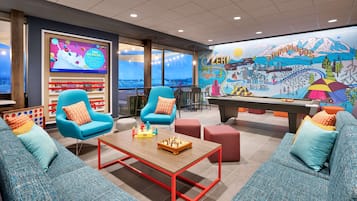 Uma área de estar moderna com dois poltronas azuis, uma mesa de centro de madeira e mesa de bilhar. Uma tela grande exibe um canal de notícias, e um mural colorido retrata uma paisagem montanhosa.