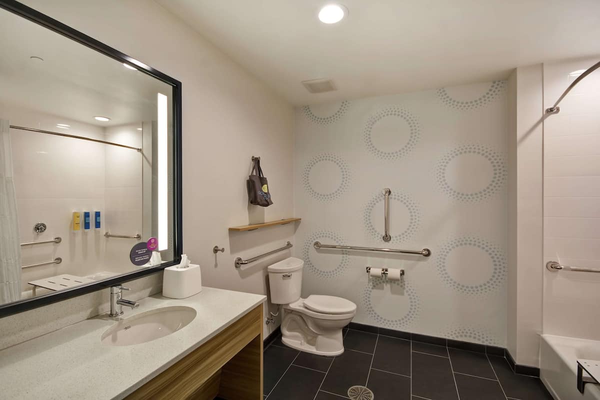 A modern bathroom with a sink, toilet, and shower. There are handrails, a mirror, and a towel rack.