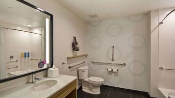 Un baño moderno con lavamanos, inodoro y ducha. Cuenta con barras de apoyo, un espejo y un perchero para toallas.