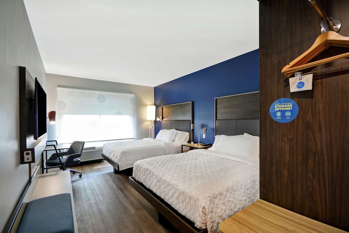 A hotel room with two beds, a desk, a chair, a TV, and a window.