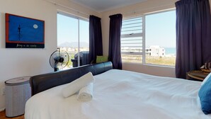 A hotel room with a bed, a fan, a window with curtains, a view of the sea, and a painting on the wall. - Copper Den Condo (Cape Town)