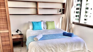 Ein ordentlich bezogenes Bett mit blauer Decke und gerolltem Handtuch, ein Nachttischchen mit Lampe und ein Fenster mit Vorhängen.