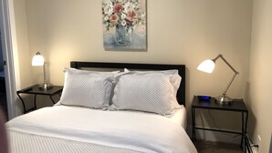Una cama bien tendida con almohadones estampados, dos lámparas de noche, un reloj y un cuadro floral en la pared.