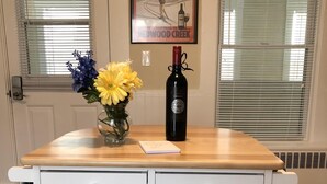 Una cocina con una mesa de madera, dos taburetes blancos, un florero con flores y una botella de vino.