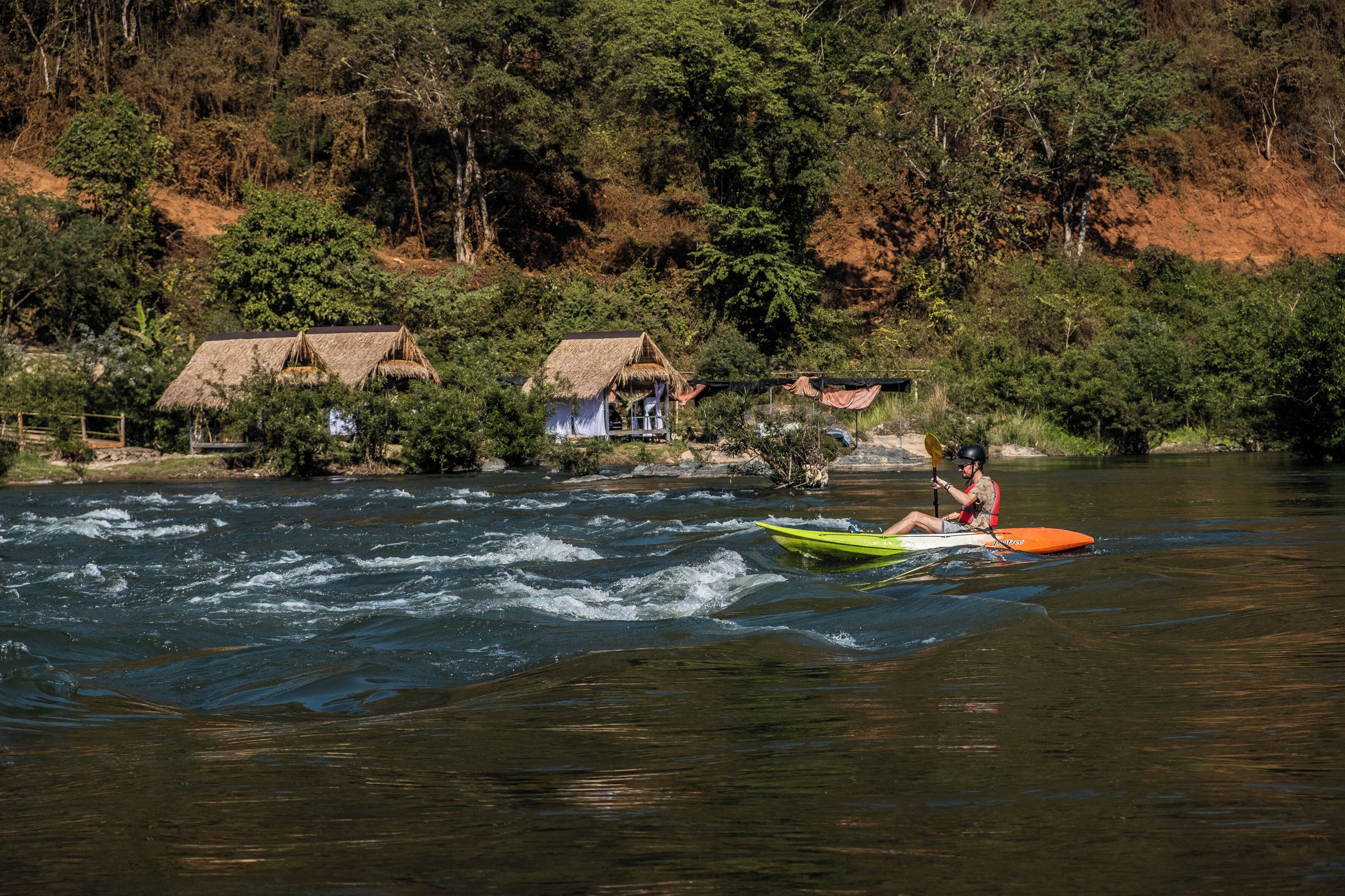 A river with a person kayaking. There are thatched-roof huts in the background.