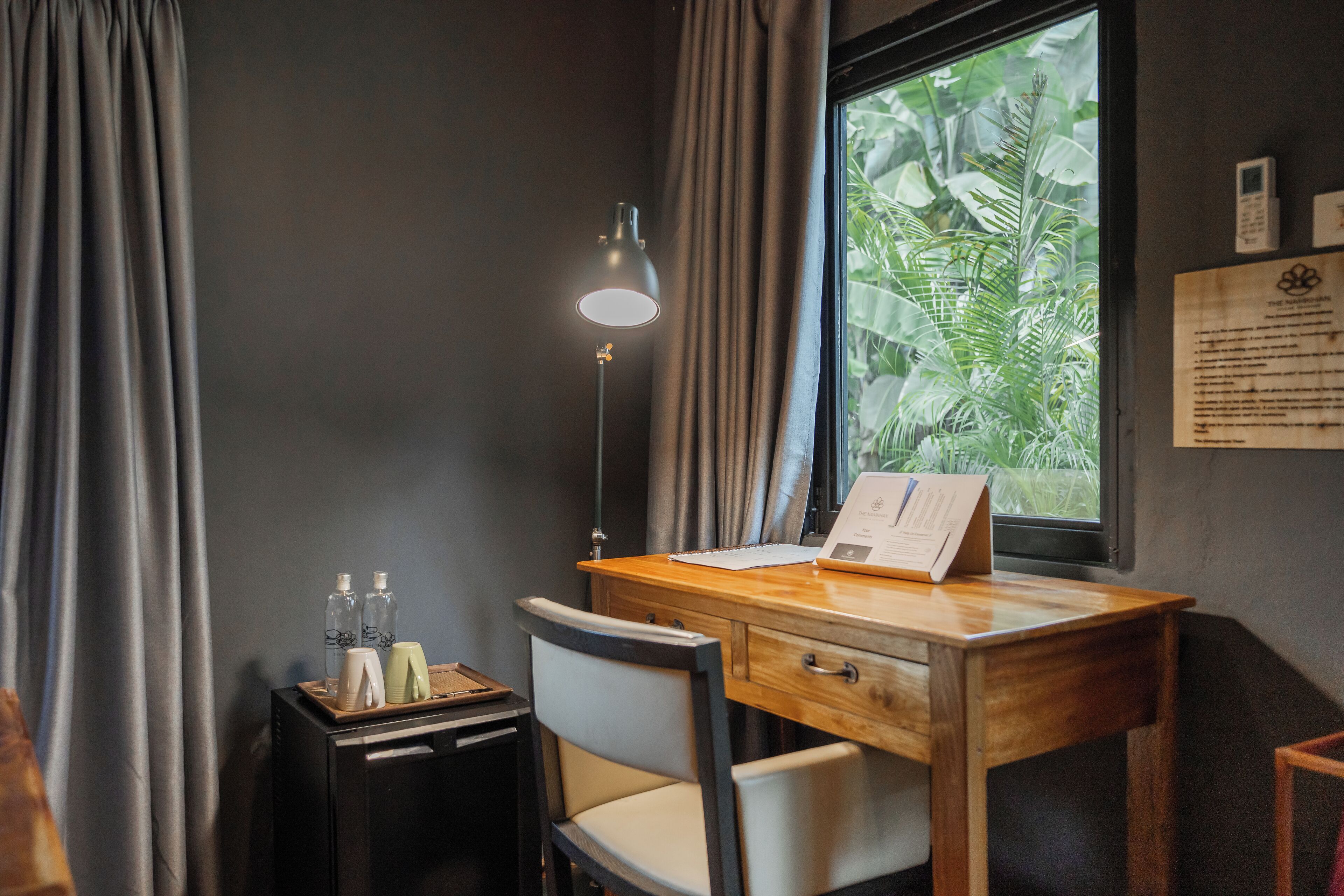 Una habitación con un escritorio de madera, una silla, una lámpara, un ventanal con vistas a la vegetación y una mesita con botellas y una libreta.