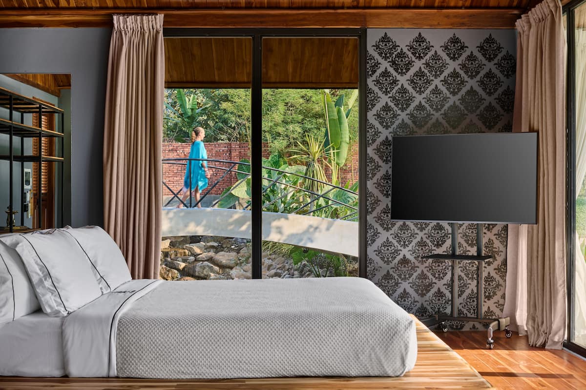 Une chambre avec un grand lit, un téléviseur posé sur un meuble, une bibliothèque et une vue sur un jardin et un pont à l’extérieur.