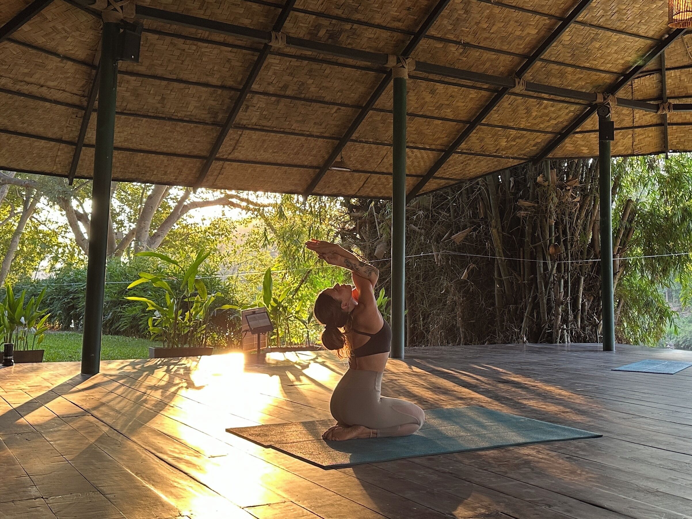 Una esterilla de yoga sobre una terraza de madera bajo un techo de paja, rodeada de follaje.