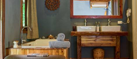 Un baño con tocador de madera, dos lavamanos, un espejo, toallero, un taburete pequeño y pantuflas en el suelo.
